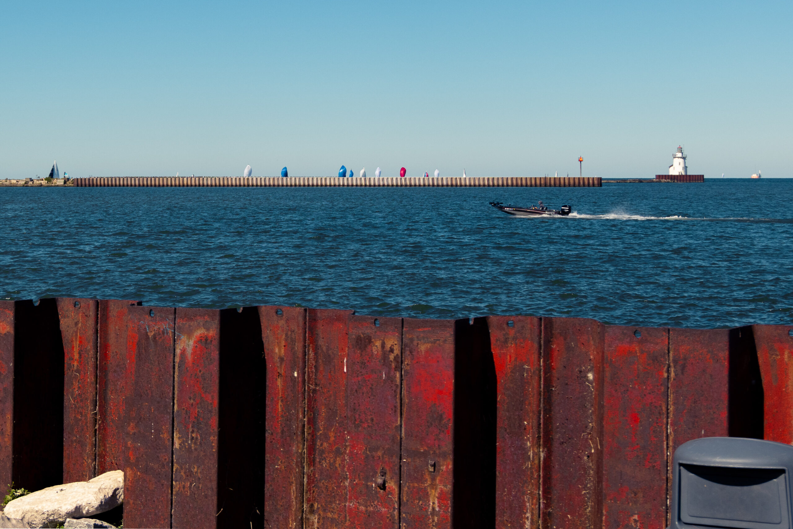 A speedboat zooms by in front of a lighthouse and a bunch of sailboats in the background. In the foreground is this weird kinda rusty red fence and the top of a trash can.