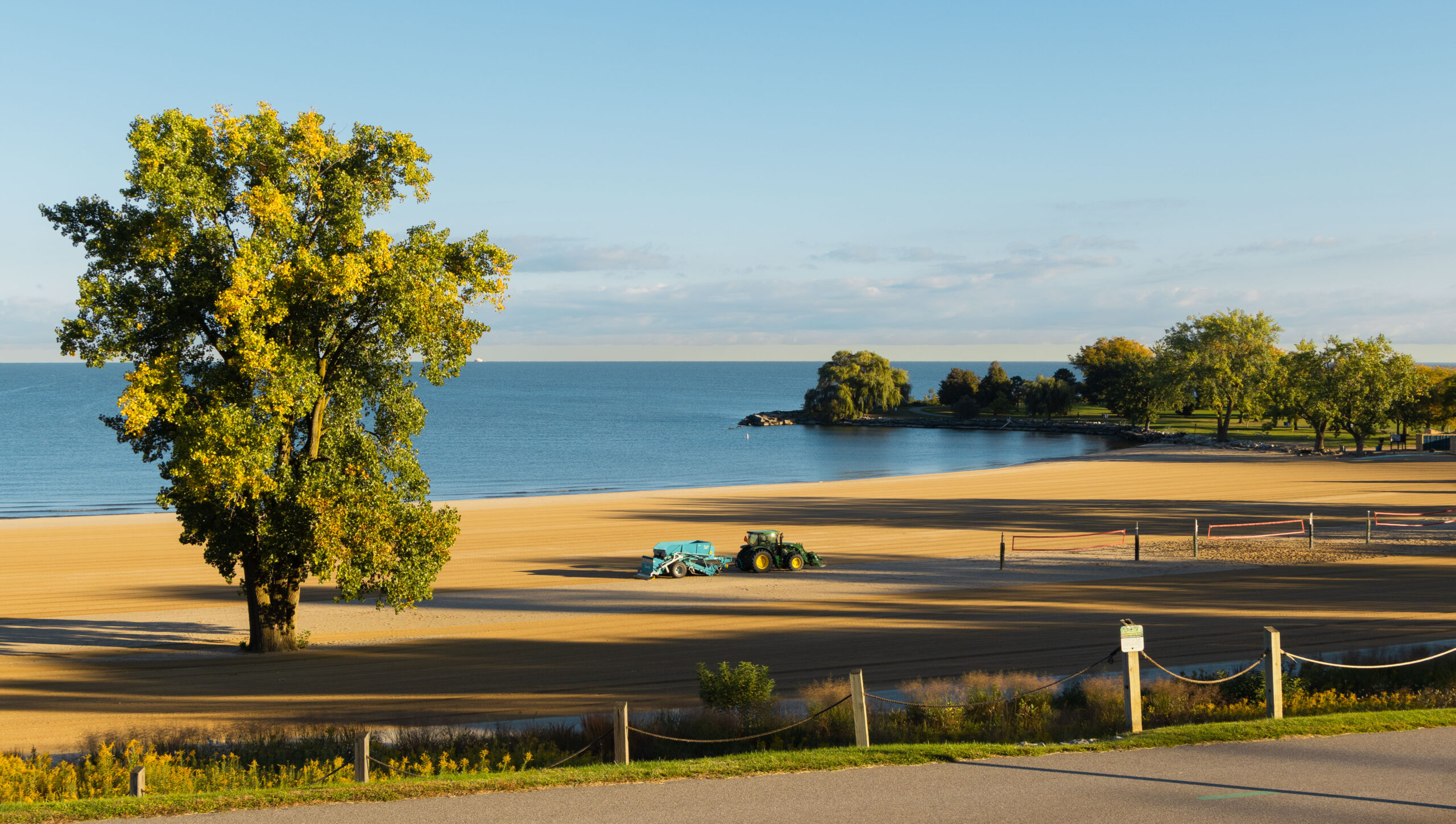 A John Deere pulls a big teal thing behind it on a wide open Edgewater Beach in Cleveland Ohio. The sun has just come up so the big tree on the beach behind it is showered in gold light.